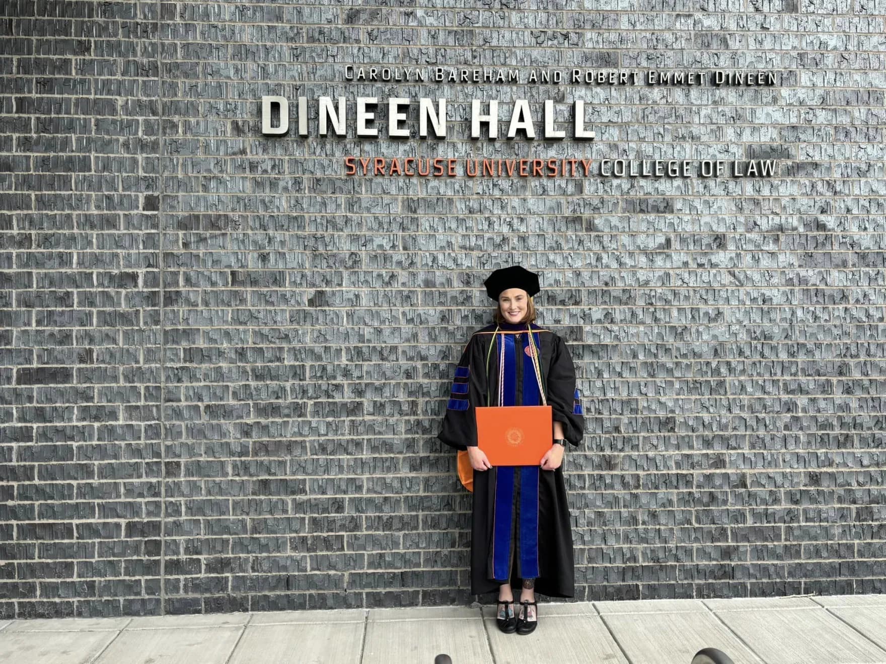Cody Nagle in cap and gown outside Dineen Hall, Syracuse University College of Law, holding her diploma